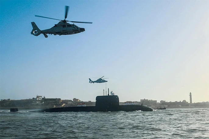 A submarine underway with helicopters flying above the coastline.