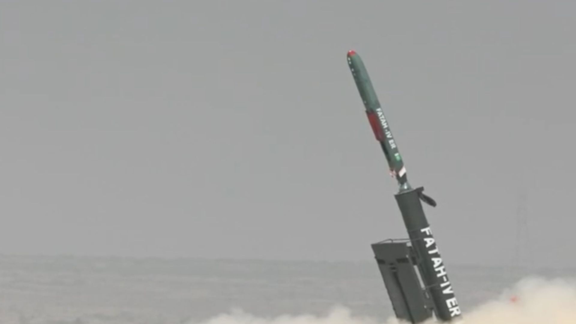 Rocket booster launching from a platform at sea, tilting upward with exhaust plume and dust surrounding it.