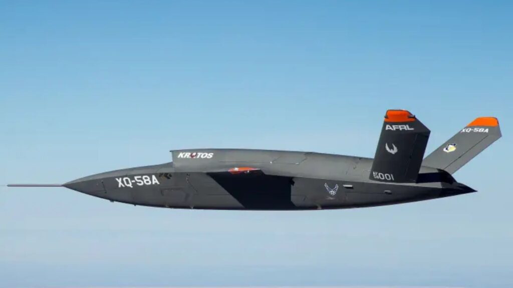 Grey XQ-58A Valkyrie unmanned combat aircraft in flight with orange-tipped tails and Kratos branding against a clear blue sky