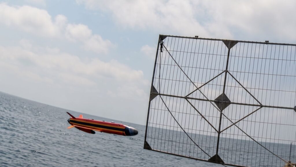 A red, black, and yellow speedboat gliding over choppy sea next to a large metal lattice fence by the water.