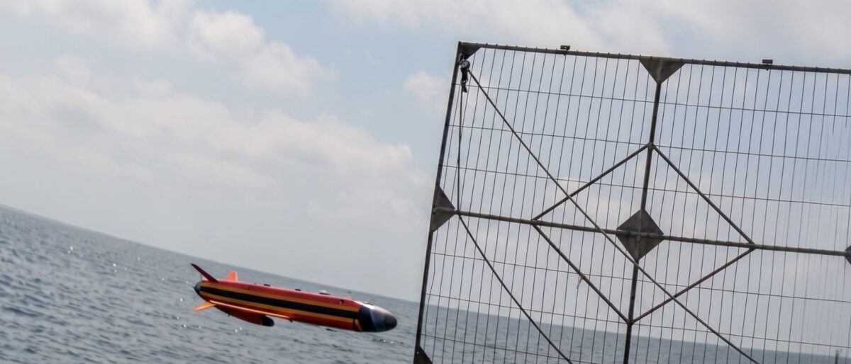 A red, black, and yellow speedboat gliding over choppy sea next to a large metal lattice fence by the water.