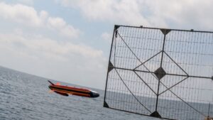 A red, black, and yellow speedboat gliding over choppy sea next to a large metal lattice fence by the water.