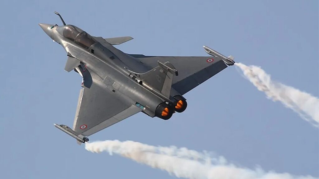 Silver military fighter jet performing a steep turn with orange afterburners and a white smoke trail against a clear blue sky, mid-air show.