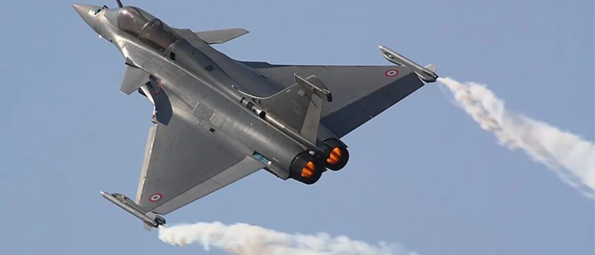 Silver military fighter jet performing a steep turn with orange afterburners and a white smoke trail against a clear blue sky, mid-air show.