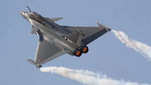 Silver military fighter jet performing a steep turn with orange afterburners and a white smoke trail against a clear blue sky, mid-air show.