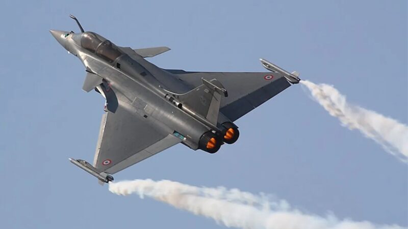 Silver military fighter jet performing a steep turn with orange afterburners and a white smoke trail against a clear blue sky, mid-air show.