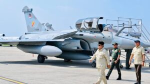 Ground crew and pilots walk beside a parked fighter jet on a tarmac, backlit by a clear sky.