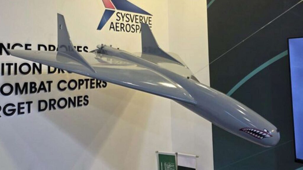 Large grey model jet displayed indoors with a company banner and product list in the background (drones, combat helicopters).