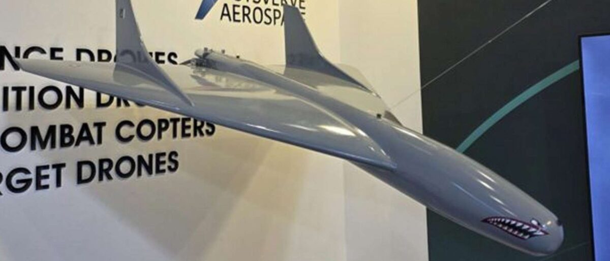 Large grey model jet displayed indoors with a company banner and product list in the background (drones, combat helicopters).
