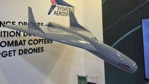 Large grey model jet displayed indoors with a company banner and product list in the background (drones, combat helicopters).