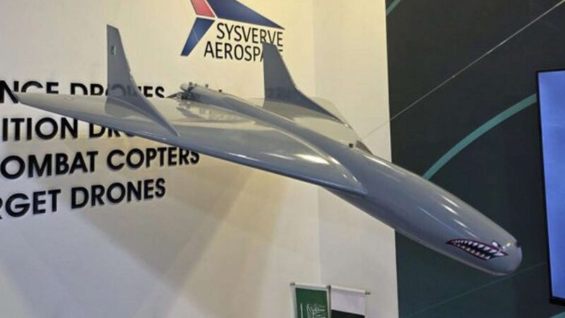 Large grey model jet displayed indoors with a company banner and product list in the background (drones, combat helicopters).