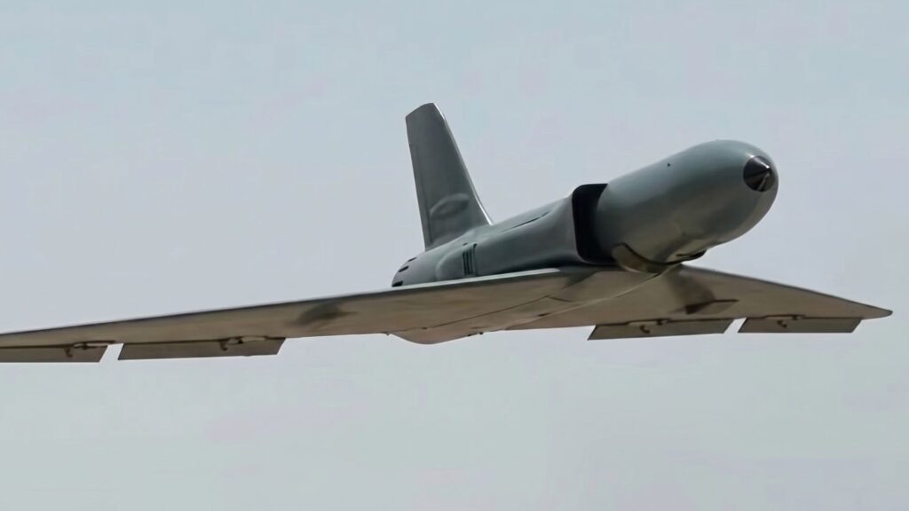 Unmanned drone in flight with a wide, flat wing and a bulbous forward fuselage/nose pod against a pale sky.