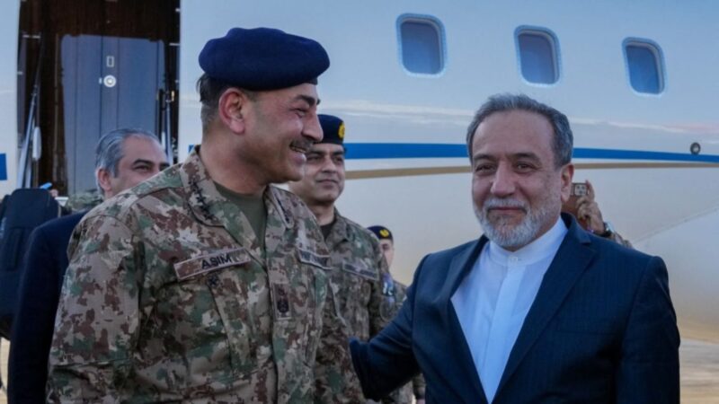 Military officer in camouflage with a blue beret smiling beside a civilian man near a plane.