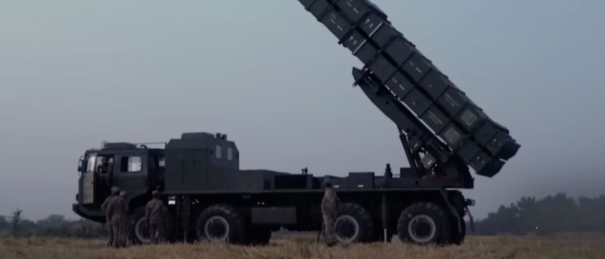 Military truck-mounted rocket launcher elevated at a steep angle with soldiers standing nearby in a field.