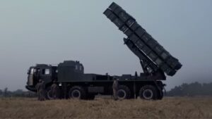 Military truck-mounted rocket launcher elevated at a steep angle with soldiers standing nearby in a field.
