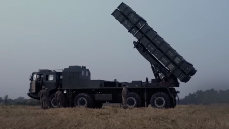 Military truck-mounted rocket launcher elevated at a steep angle with soldiers standing nearby in a field.
