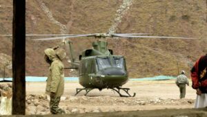 Green military helicopter on a dirt pad with rotor blades spinning and personnel nearby in a rocky, mountainous base setting.
