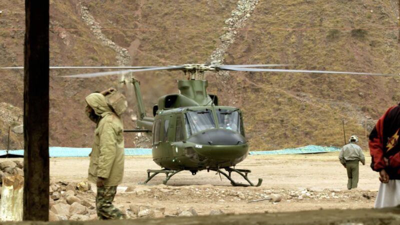 Green military helicopter on a dirt pad with rotor blades spinning and personnel nearby in a rocky, mountainous base setting.