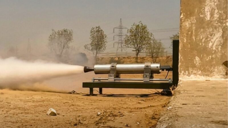 Rocket motor firing on a test stand in a dusty desert site, exhaust plume blowing to the left.