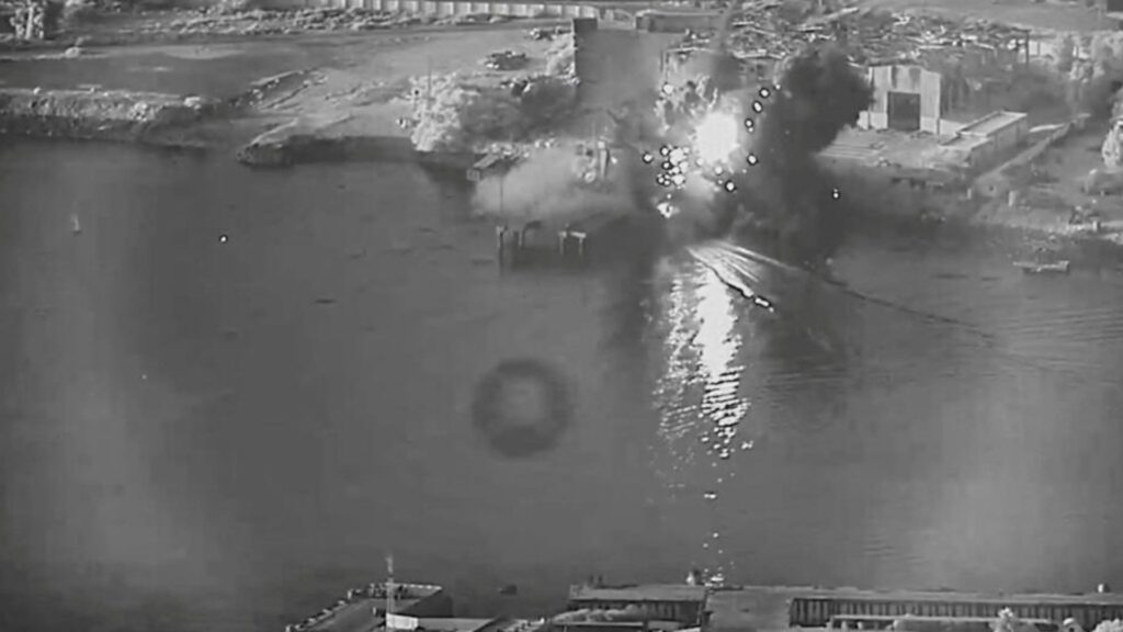 Harbor explosion: a bright fireball and smoke plume rising from the water near industrial buildings and a quay.