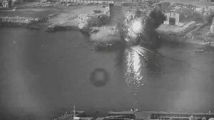 Harbor explosion: a bright fireball and smoke plume rising from the water near industrial buildings and a quay.