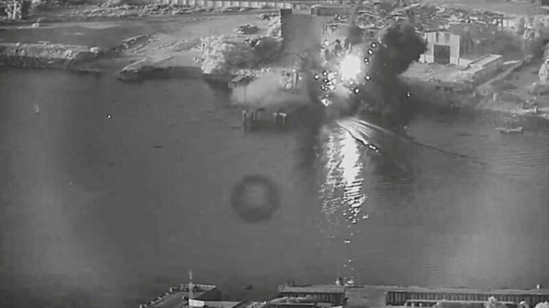 Harbor explosion: a bright fireball and smoke plume rising from the water near industrial buildings and a quay.