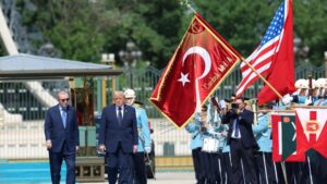 Two suited men walking past a ceremonial military parade with American and Turkish flags in the background.