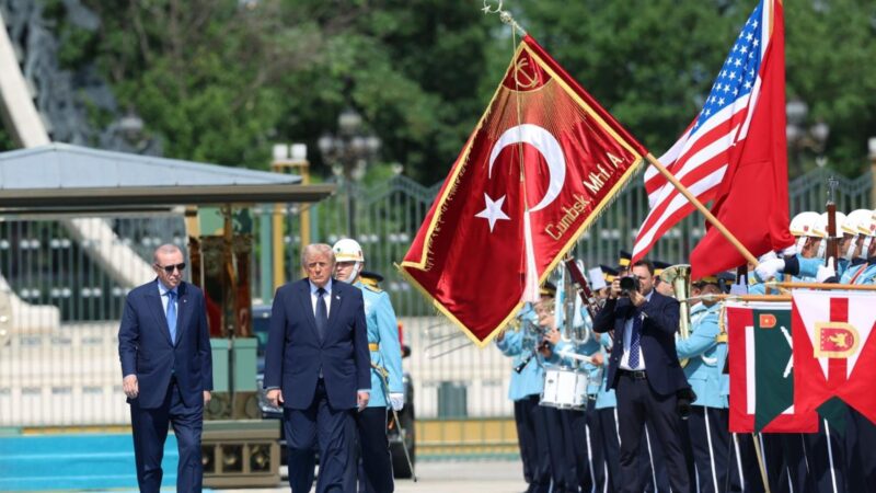 Two suited men walking past a ceremonial military parade with American and Turkish flags in the background.