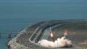 Rocket-style flame and white smoke erupting from a ground test site along a curved breakwater by the sea.