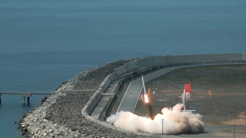 Rocket-style flame and white smoke erupting from a ground test site along a curved breakwater by the sea.