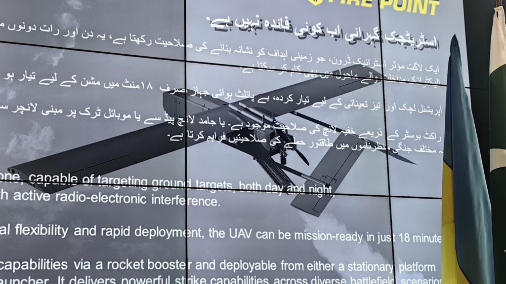 Large video wall showing multiline text in Urdu/Arabic and English about UAV capabilities, with a plane wing image and clouds in the background.