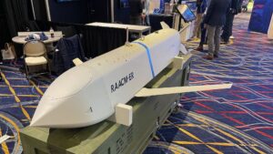 White unmanned aerial vehicle fuselage on a wheeled cart at a trade show, with 'RAACMER-ER' printed on its side and people in business attire in the background.