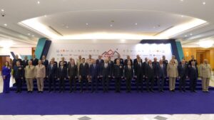 Formal group photo of military officers and officials in suits on a blue-carpeted stage at a conference, backdrop with logos behind them.