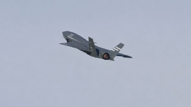 A gray military fighter jet in flight with afterburner glowing orange, angled slightly upward against a pale blue sky.
