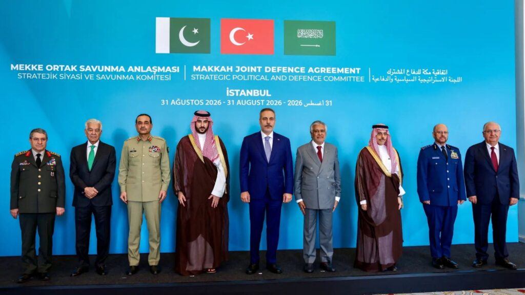 Group of eight men in military uniforms and suits standing in a line on a stage with a bright blue backdrop featuring flags and the title 'Makkah Joint Defence Agreement' and related text.