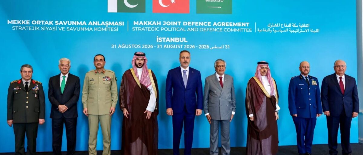 Group of eight men in military uniforms and suits standing in a line on a stage with a bright blue backdrop featuring flags and the title 'Makkah Joint Defence Agreement' and related text.