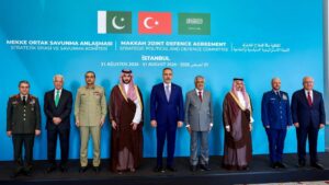 Group of eight men in military uniforms and suits standing in a line on a stage with a bright blue backdrop featuring flags and the title 'Makkah Joint Defence Agreement' and related text.