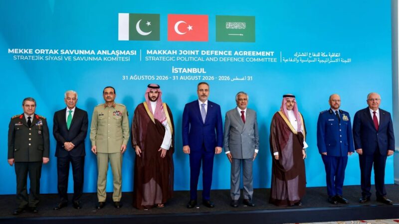 Group of eight men in military uniforms and suits standing in a line on a stage with a bright blue backdrop featuring flags and the title 'Makkah Joint Defence Agreement' and related text.