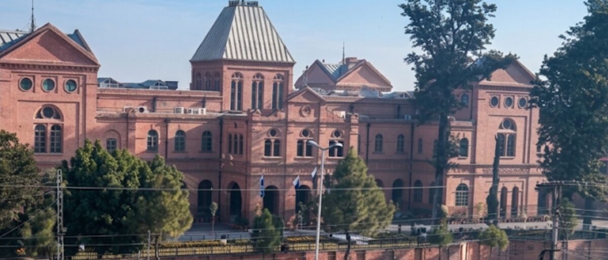 Red-brick university building with arched windows and a tall central roof, surrounded by trees on a sunny day