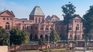 Red-brick university building with arched windows and a tall central roof, surrounded by trees on a sunny day