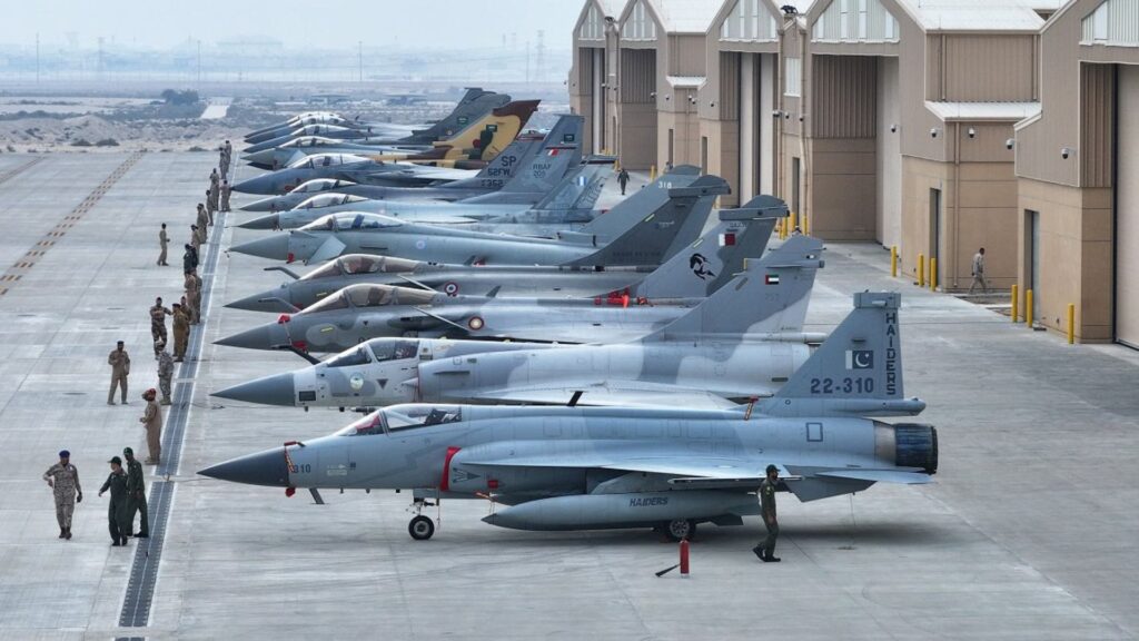 Row of fighter jets parked on a concrete tarmac beside hangars, with personnel walking along.