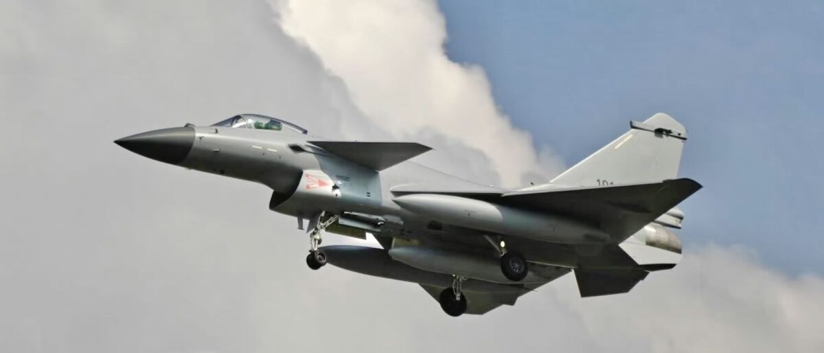 Fighter jet taking off with landing gear down, gray aircraft against a blue sky with clouds behind it.