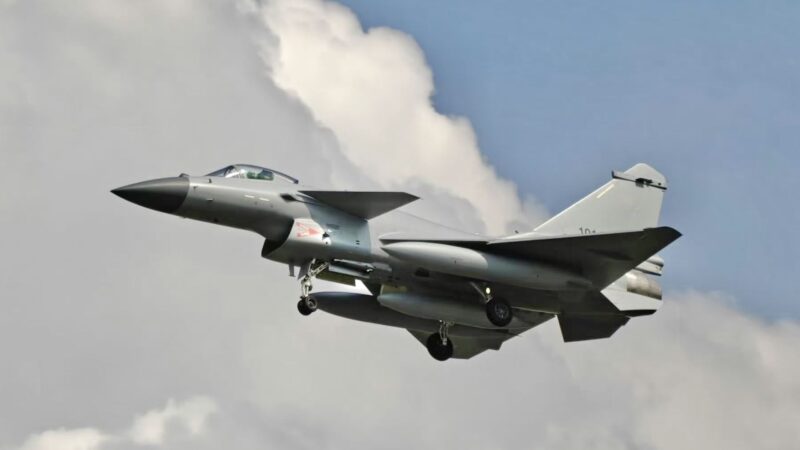 Fighter jet taking off with landing gear down, gray aircraft against a blue sky with clouds behind it.