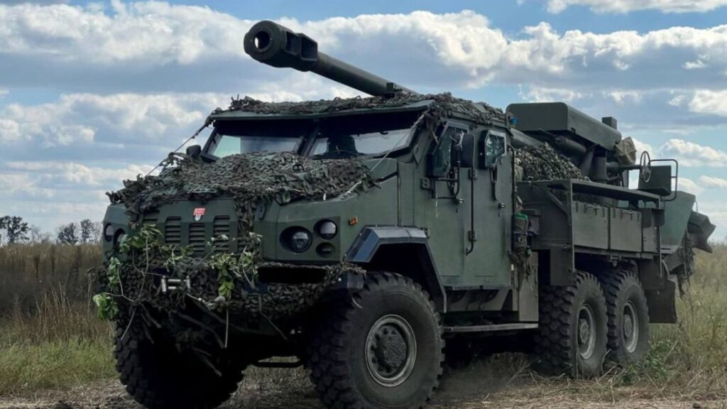 Armored self-propelled artillery vehicle with a long gun, camouflaged with netting in an open field under a blue sky.