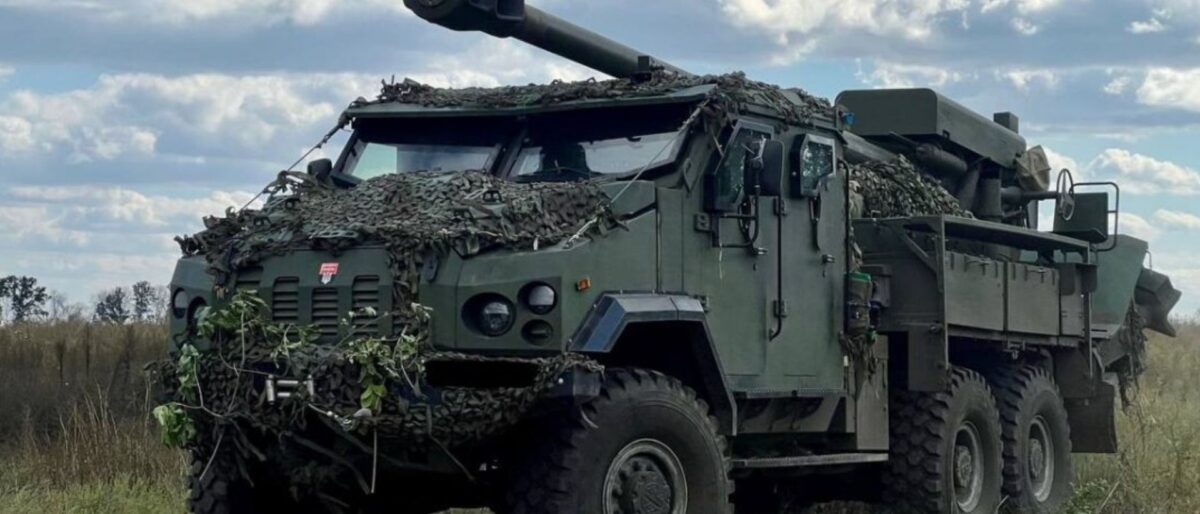 Armored self-propelled artillery vehicle with a long gun, camouflaged with netting in an open field under a blue sky.