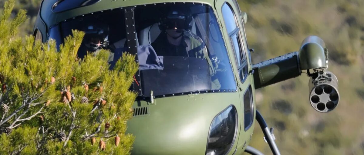 Olive-green military helicopter flying low among pine branches, with a side rocket launcher visible on its arm.