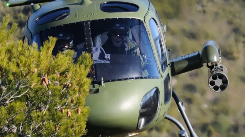 Olive-green military helicopter flying low among pine branches, with a side rocket launcher visible on its arm.
