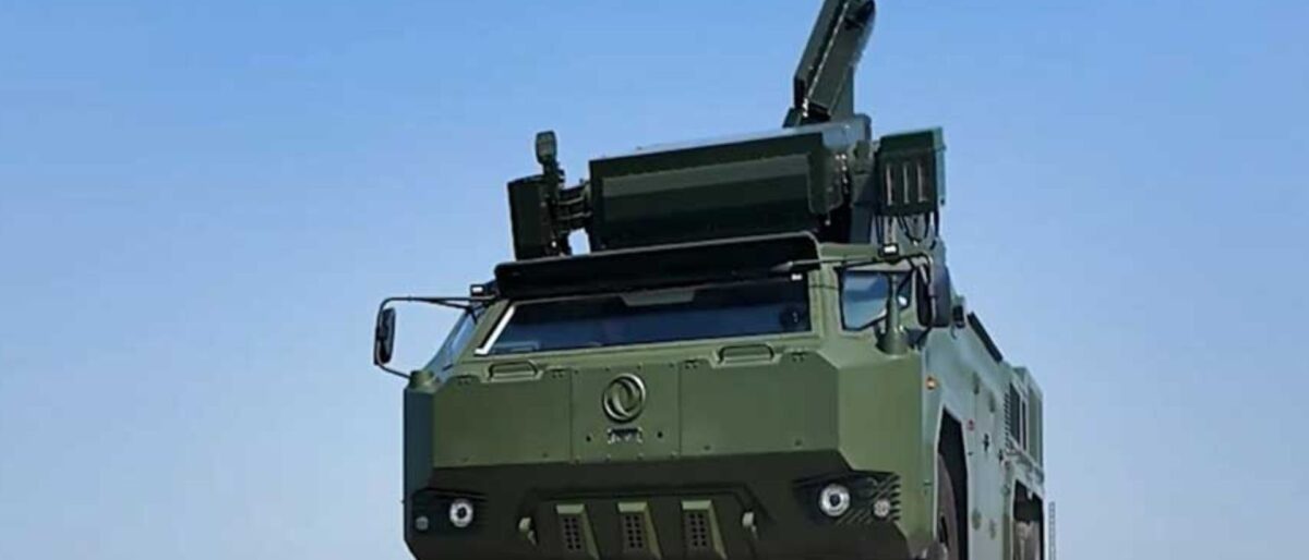 Green armored military truck with an elevated rocket launcher on top against a clear blue sky.