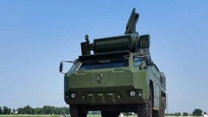 Green armored military truck with an elevated rocket launcher on top against a clear blue sky.