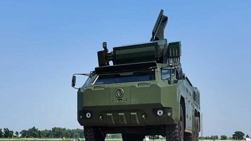 Green armored military truck with an elevated rocket launcher on top against a clear blue sky.